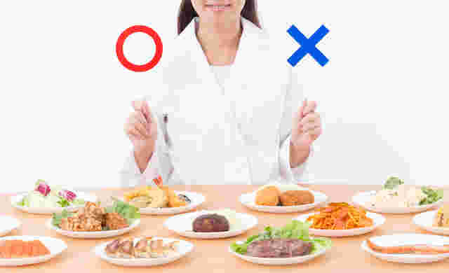 白衣を着た女性がマルとバツの棒を持ち、小さめのお皿に色んな食材が乗せてある