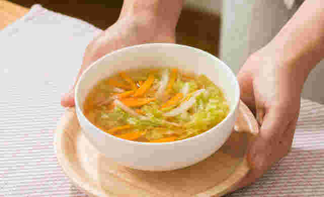 木の小さめのおぼんに丸い茶碗の中にカットされた野菜とスープが入っているのを両手で持っている