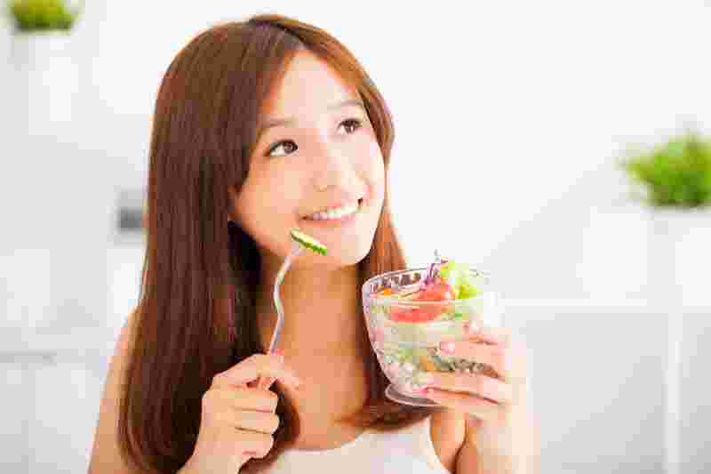 野菜の入ったガラスの器を手に持ちきゅうりのスライスを口に運ぶ笑顔の女性