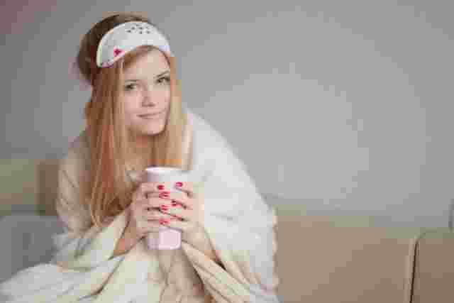 毛布にくるまりアイマスクをおでこにつけて、カップを両手に持った女性