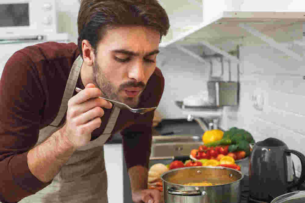キッチンで外国人男性が手にスプーン持ち、味を確かめている様子