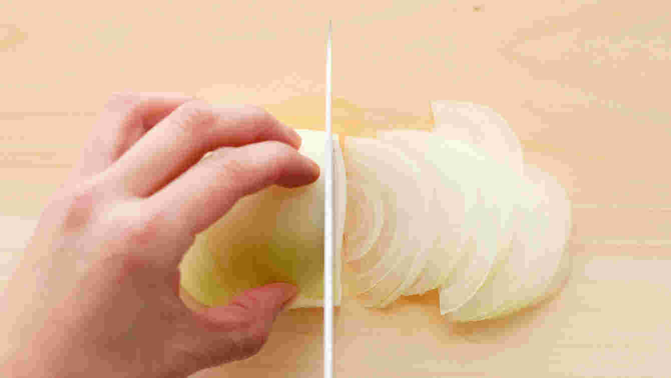 木のまな板の上に皮をむいた半玉ねぎに手をそえて包丁で切っている