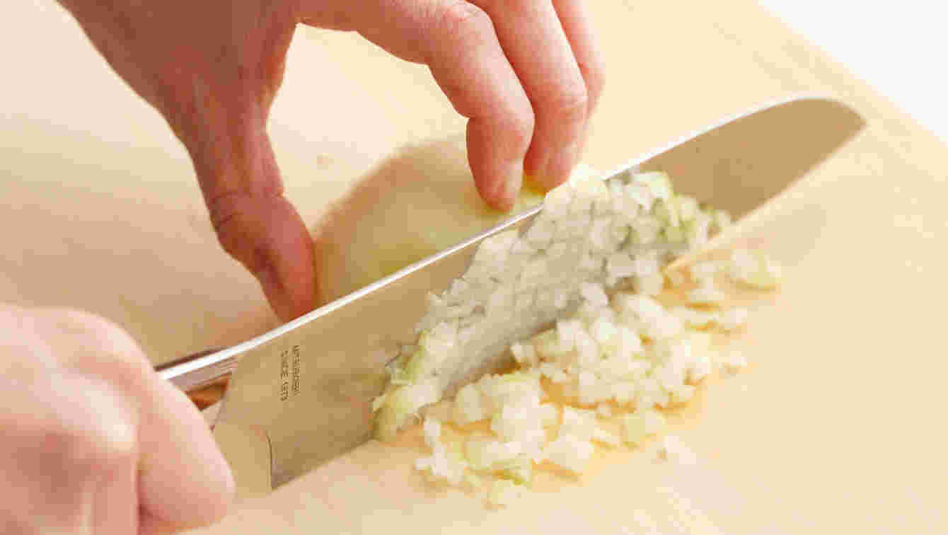 木のまな板の上に皮をむいた半玉ねぎに手をそえて包丁でみじん切りにしている