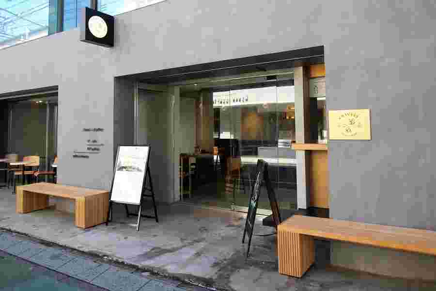 グレーの壁で木製のベンチを2つ外に出しているオシャレなお店の外観