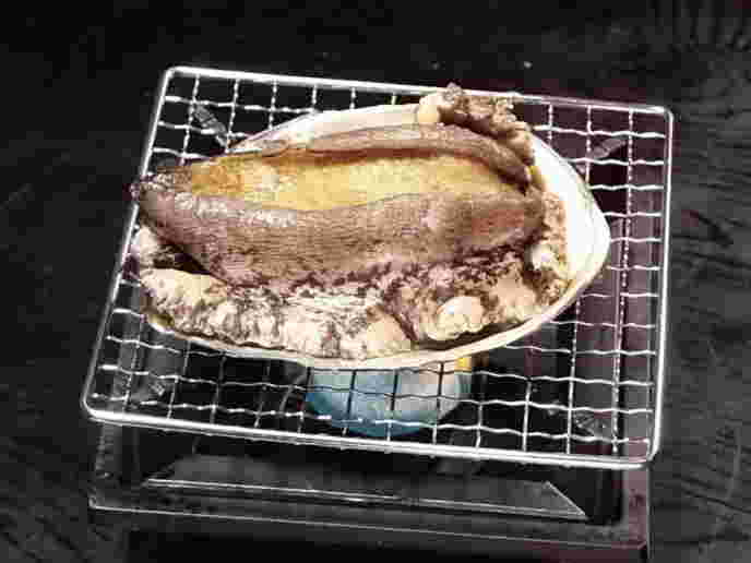 カセットコンロの上に網が引かれ、1個のアワビが裏返され置かれている。